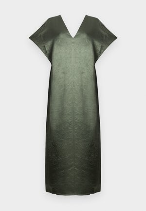 Kvinde står mod hvid baggrund iført en silkeagtig grøn midi-kjole med V-hals, bærer en sort skuldertaske og beige spidse sko.