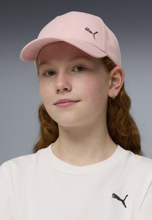 Joven de piel clara y cabello rizado y rojo que lleva una gorra Puma rosa claro y una camiseta blanca de Puma sobre un fondo gris.
