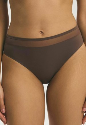 Kobieta w brązowych majtkach bikini z przezroczystym pasem, ukazująca brzuch i górną część ud na neutralnym tle.