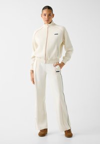 Frau steht in einem cremefarbenen Guess-Trainingsanzug mit Reißverschlussjacke und passenden Hosen, kombiniert mit braunen Wildlederschuhen, vor weißem Hintergrund.