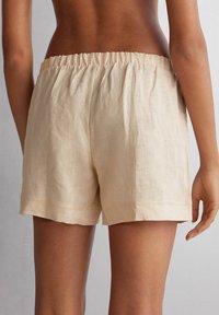 Pantalones cortos de lino beige con cintura elástica, que presentan un corte holgado y una textura sutil. Piernas dobladadas sin cierres o acentos visibles.