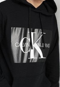 Felpa nera con cappuccio caratterizzata da una grande grafica rettangolare lucida che presenta "CK" e "CALVIN KLEIN JEANS" in bold bianco. Stile pullover con cordini.