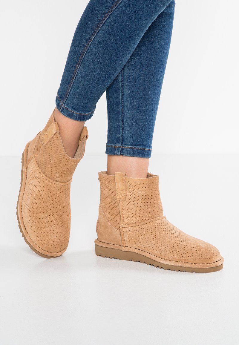 Bottes cheville en daim beige avec design perforé, semelle en caoutchouc plate et languette à l'arrière. Portées avec un jean bleu, affichant une coupe décontractée.