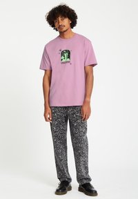 Lichtpaarse T-shirt met een zwart grafisch ontwerp van bomen en sterren. Gecombineerd met zwarte broek die een wit abstract patroon toont.