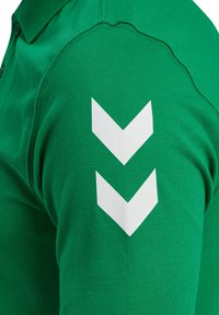 Grön pikétröja med två vita nedåtvända chevronränder nära axels�ömmen, som visar tygstruktur och sömdetaljer.