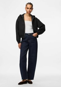 Felpa nera con zip e orlo a coste, indossata sopra un top bianco. I jeans a gamba larga blu scuro e le scarpe flats marroni completano l'outfit.