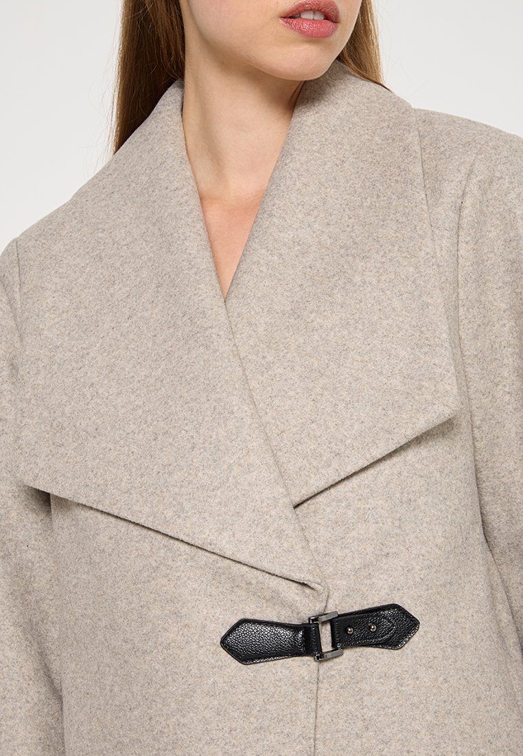 Abrigo de lana gris claro con solapa ancha, tejido texturizado y cierre de cinturón de cuero negro con un acento metálico plateado.