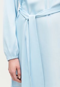 Χέρι που φοράει ανοιχτό φυσικό βερνίκι νυχιών δίπλα σε ανοιχτό μπλε φόρεμα με μακριά μανίκια και ζώνη στη μέση.