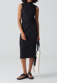 Robe sans manches noire texturée avec un col haut et une coupe à mi-genoux. Associée à des sandales noires et un sac fourre-tout blanc avec un accent noir.