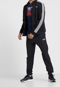 Czarna kurtka dresowa z białymi paskami, w połączeniu z czarnymi spodniami z logo. Buty są czarne z białą podeszwą. Materiał wydaje się lekki.