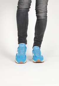 Blue athletic shoes made of suede and mesh, featuring white accents, a textured sole, and flat laces, worn with dark gray jeans.