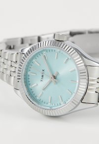 Silver Timex watch with a round, teal face, silver hour markers, and a textured bezel. Features a stainless steel bracelet.