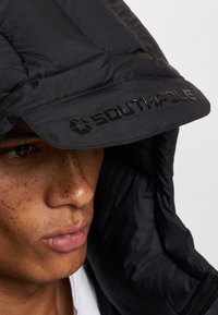 Un bonnet en duvet noir avec un logo "SOUTHPole" noir brillant sur le visière. Texture lisse, gros plan sur les traits du visage avec un fond neutre.