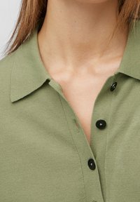 Gros plan d'une personne portant une chemise verte à boutons avec col, trois boutons noirs visibles, et des cheveux châtain clair tombant sur les épaules.