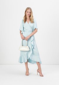 Vestido envolvente azul claro com mangas curtas e bainha com babados, combinado com uma bolsa branca e saltos nu com tiras. Textura suave em todo o vestido.