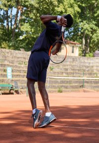 Αθλητής σε ναυτικές αθλητικές ενδυμασίες σε χωμάτινη επιφάνεια τένις, έτοιμος να σερβίρει. Γκρίζα τενιστικά παπούτσια με λευκές λεπτομέρειες, δυναμική στάση, ρακέτα στο χέρι.