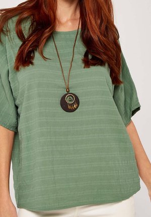 Vrouw met lang rood haar, die een losse groene getextureerde top draagt en een ronde hanger ketting met hangende bedels aan een bruine koord.