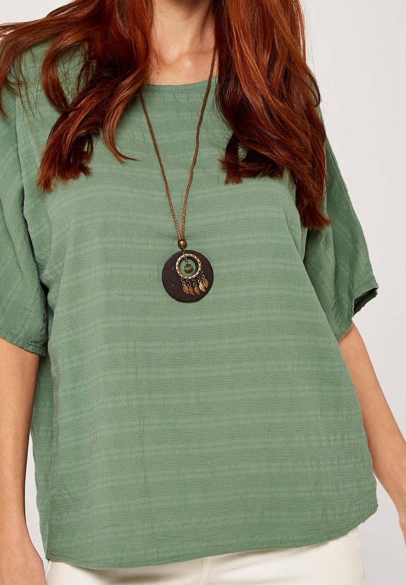 Femme aux longs cheveux roux portant un haut vert texturé ample et un collier pendentif rond avec des breloques suspendues sur un cordon marron.