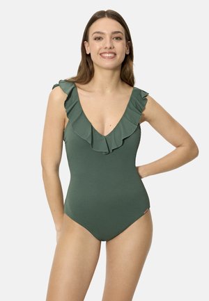 Joven sonriente, con un traje de baño entero verde de escote en V profundo con volantes, apoyando su mano derecha en la cadera.