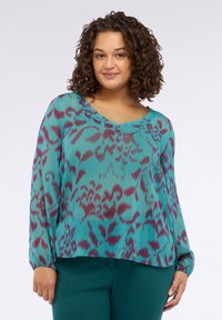 Donna che indossa una blusa a maniche lunghe turchese con stampa floreale astratta viola e pantaloni verde scuro, in piedi su uno sfondo bianco semplice.