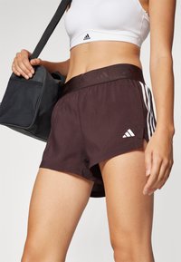 Shorts de sport marron avec des accents blancs à trois rayures, une large ceinture et une coupe ample. Le matériau semble léger et lisse.