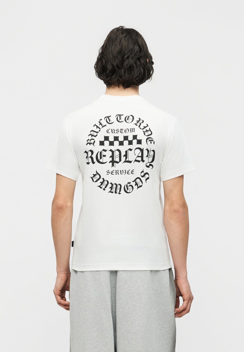 Άτομο με κοντά σκούρα μαλλιά που φοράει λευκό T-shirt με μαύρο γοτθικό κείμενο και γραφικό με σκακιέρα στην πλάτη, γκρι παντελόνι.