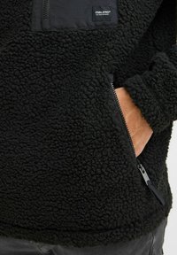 Black fleece pullover with a textured, fuzzy surface, chest pocket in contrasting material, and side zipper. Includes a hand pocket.