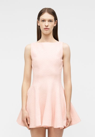 Γυναίκα με καστανά ίσια μαλλιά που φοράει ροζ αμάνικο φόρεμα με φούστα που ανοίγει, στέκεται μπροστά σε απλό ανοιχτόχρωμο φόντο.