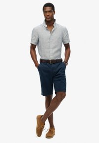 Camisa de rayas azul claro de mangas cortas, pantalones cortos azul marino, zapatos de ante marrón, cinturón marrón. Corte clásico, estilo casual, acentos mínimos.
