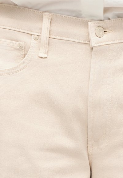 Εγγύς λήψη μπεζ τζιν υφάσματος, που δείχνει τη μέση, το κουμπί, τη θηλιά της ζώνης, την μπροστινή τσέπη και τις ορατές λεπτομέρειες των ραφών.