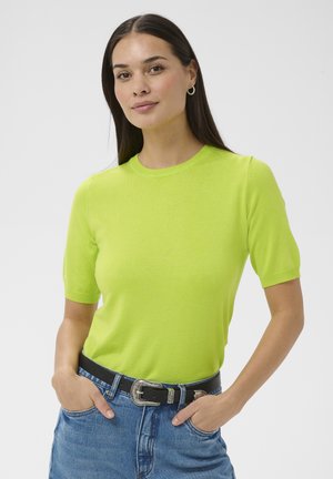 Femme portant un haut à manches courtes vert vif, un jean bleu avec une ceinture noire, mains dans les poches, et de petites créoles, sur un fond blanc.