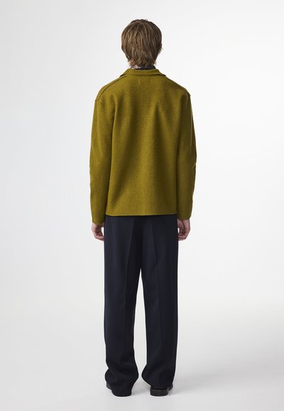 Giacca in lana verde, con una texture ed una vestibilità ampia, cuciture visibili e colletto, abbinata a pantaloni scuri, sartoriali.