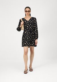 Μαύρο φόρεμα με v-λαιμό, μακριές φούσκες μανίκια και ζώνη που δένει. Διαθέτει μπεζ αφηρημένο τετραγωνικό μοτίβο. Συνδυάζεται με μαύρες σανδάλες με λουράκια.