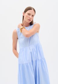 Ljusblå ärmlös klänning i mjukt tyg, med en lagerdesign och en flödande siluett. Modellen bär ett guldarmband på sin arm.