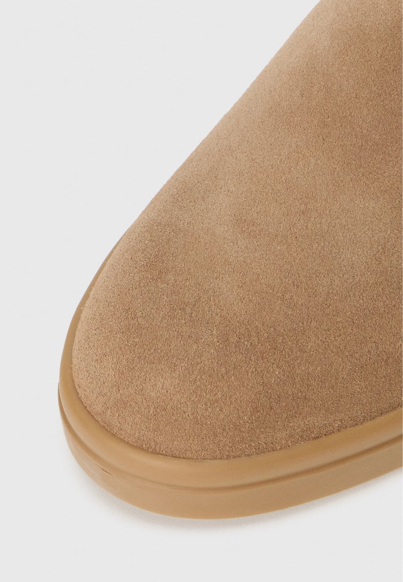 Primo piano di una scarpa in suede color tan, caratterizzata da una texture morbida e una punta arrotondata, con una suola in gomma beige per il contrasto.