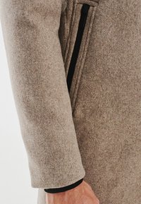 Beżowy wełniany płaszcz o teksturowanym wykończeniu, z czarną kieszenią na zamek błyskawiczny po boku. Rękaw ma czysty, dopasowany krój.