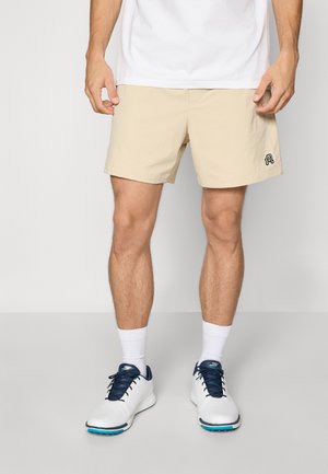 Men's beige shorts made of a lightweight material, featuring a black logo on the left leg, paired with white socks and navy-trimmed sneakers.