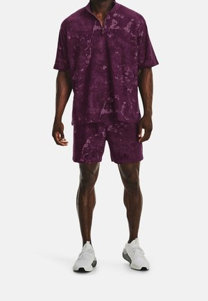 Mann trägt ein lila kurzärmeliges Zip-Up-Shirt und passende Shorts mit weißen Sportschuhen, steht vor einem weißen Hintergrund.