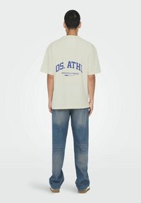 Kræmfarvet oversized T-shirt med blå tekst "OS. ATHL" og "VERSATILE HYBRIDS" på ryggen, kombineret med lyseblå jeans.