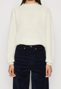 Sukienka w kolorze off-white, dziergana, z okrągłym dekoltem i prążkowaną fakturą, w połączeniu z ciemnymi spodniami z korduroju, wyposażonymi w guzik i kieszenie.
