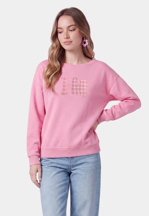 Giovane donna con capelli lunghi e mossi indossa una felpa rosa con disegno di gatto e jeans azzurri chiari, in piedi con una mano sui fianchi.