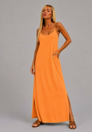 Femme aux longs cheveux blonds portant une robe maxi orange sans manches avec des fentes sur les côtés et des sandales plates marron, debout devant un fond uni.