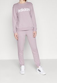 Světle fialová mikina Adidas s bílým logem, spárovaná se světlými fialovými tepláky. Oblečení vyrobené z měkké látky, s přiléhavým střihem.