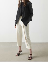 Veste en suede et cuir noir avec un devant drapé, associée à un pantalon crème ajusté avec un ourlet. Talons ouverts noirs à lanières.