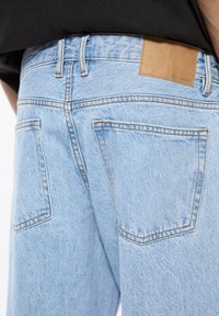Blugi din denim albastru deschis cu o textură estompată, având o patch din piele maro pe spate și buzunare posterioare cu detalii vizibile ale cusăturii.