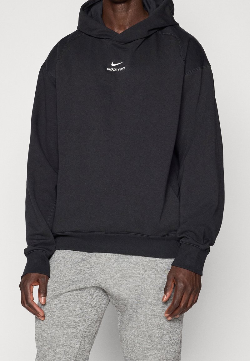Personne portant un sweat à capuche Nike Pro noir et un pantalon de survêtement gris clair, debout avec les bras détendus le long du corps sur un fond uni.