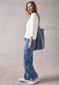 Weißes Langarmoberteil, blaue weit geschnittene Jeans und weiße Sneaker. Trägt eine graue Tote-Bag. Sanfte Texturen und lässiges Design.