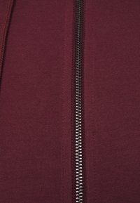 Burgunderfärgad zip-hoodie i tyg med en slät textur och synlig metalldragkedja ner i mitten. Enkelt design, inga mönster eller accenter.