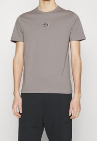 Grå bomulls-T-shirt med rund halsringning och korta ärmar. Har en svart rektangulär logotyp-patch på bröstet. Sömmar längs axlarna.