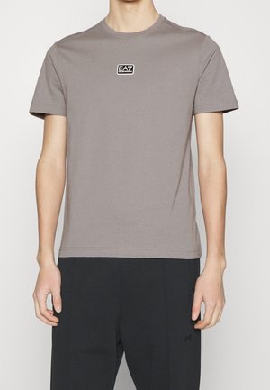 Camiseta de algodón gris con cuello redondo y mangas cortas. Presenta un parche de logo rectangular negro en el pecho. Detalle de costura en los hombros.